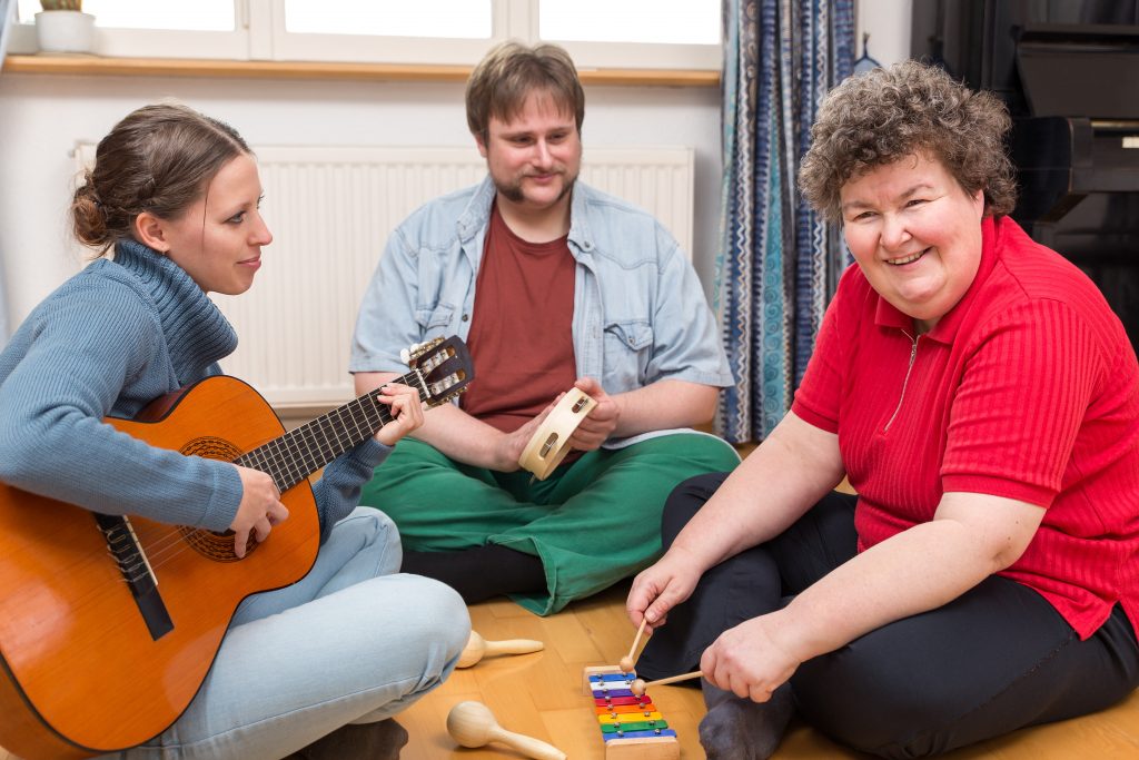A support worker playing guitar with two residents in a living room.
