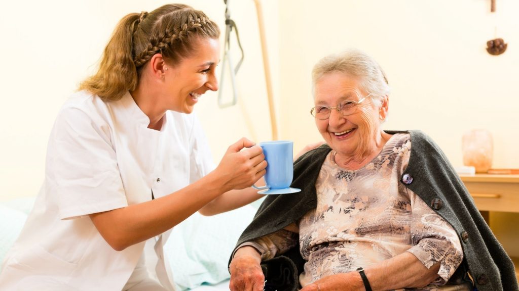 A carer smiling as she hands a cup of tea to an older woman.